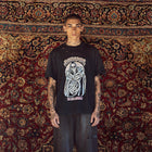 A man stands facing forward wearing a black graphic T-shirt with a large white and gray grim reaper design and the text 'TO DIE AMONG US' below it, paired with dark wide-leg jeans, in front of a large patterned red and gold rug.