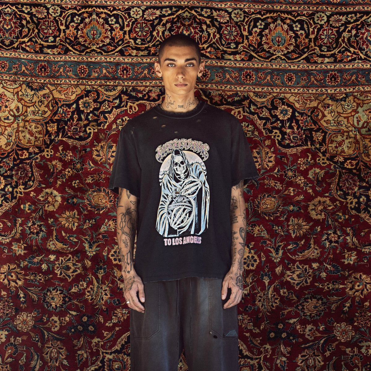 A man stands facing forward wearing a black graphic T-shirt with a large white and gray grim reaper design and the text 'TO DIE AMONG US' below it, paired with dark wide-leg jeans, in front of a large patterned red and gold rug.