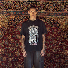 A man stands facing forward wearing a black graphic T-shirt with a large white and gray grim reaper design and the text 'TO DIE AMONG US' below it, paired with dark wide-leg jeans, in front of a large patterned red and gold rug.
