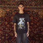 A tattooed male model stands facing forward in front of a large patterned oriental rug, wearing a black distressed Los Angeles-based graphic t-shirt and dark gray baggy jeans.