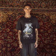 A tattooed male model stands facing forward in front of a large patterned oriental rug, wearing a black distressed Los Angeles-based graphic t-shirt and dark gray baggy jeans.