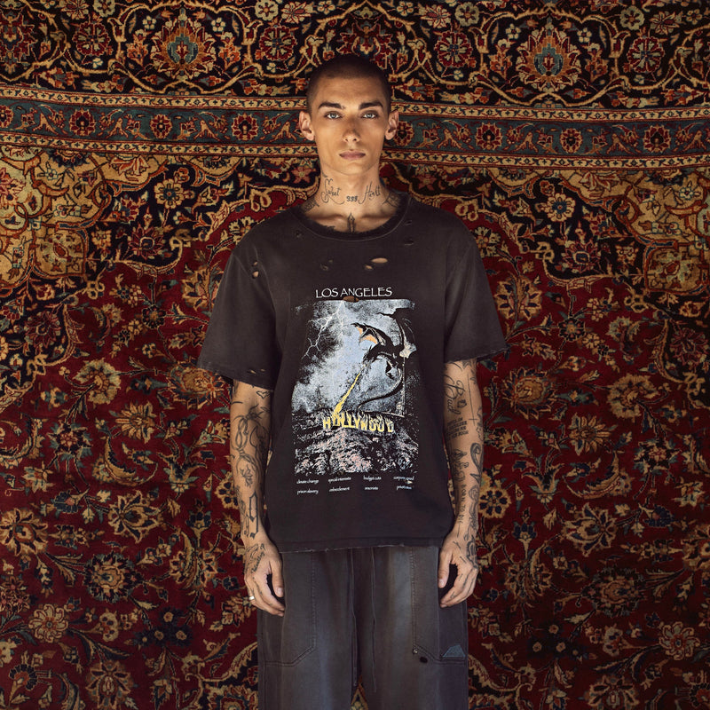 A tattooed male model stands facing forward in front of a large patterned oriental rug, wearing a black distressed Los Angeles-based graphic t-shirt and dark gray baggy jeans.