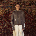 A model stands facing forward wearing a dark brown cropped hoodie with white sleeve stripes, paired with off-white wide-leg pants, posed against a large, ornate red and gold patterned rug.