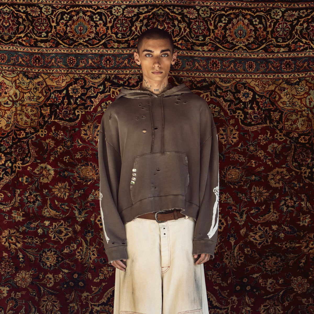 A model stands facing forward wearing a dark brown cropped hoodie with white sleeve stripes, paired with off-white wide-leg pants, posed against a large, ornate red and gold patterned rug.