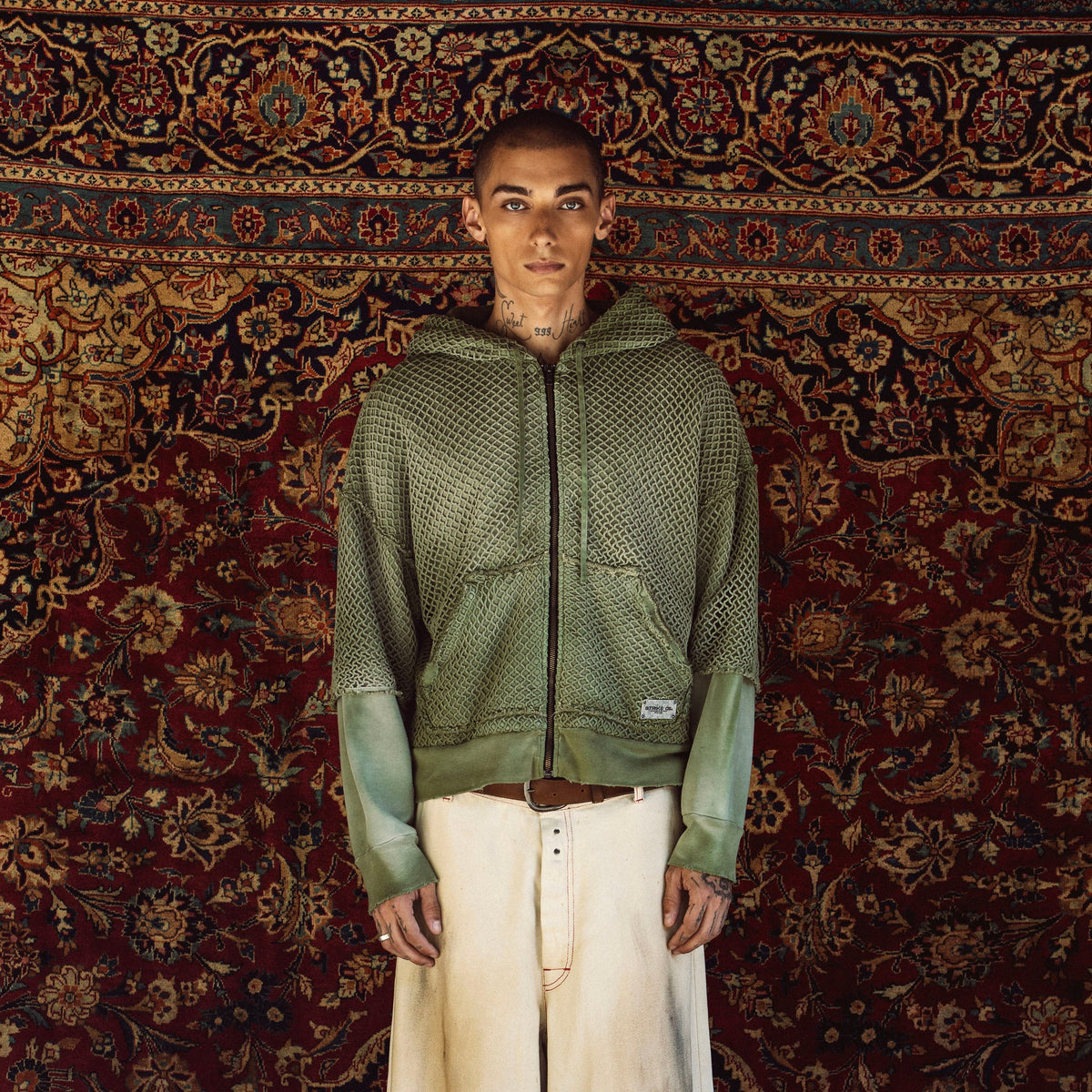 A model stands facing forward wearing a green textured zip-up hoodie with contrasting sage green sleeves, paired with off-white wide-leg jeans, against a large, ornate red and gold patterned rug.