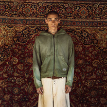 A model stands facing forward wearing a green textured zip-up hoodie with contrasting sage green sleeves, paired with off-white wide-leg jeans, against a large, ornate red and gold patterned rug.