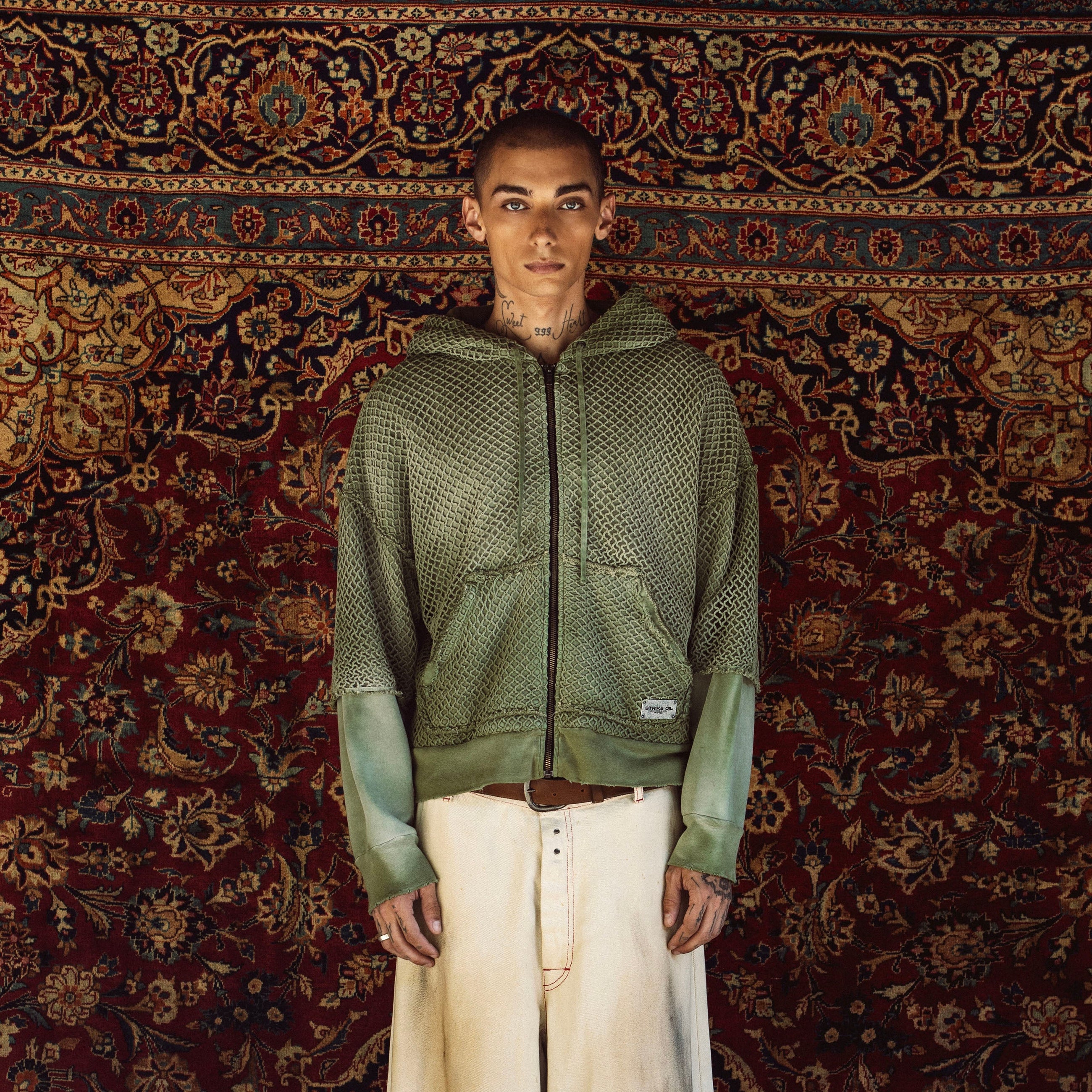 A model stands facing forward wearing a green textured zip-up hoodie with contrasting sage green sleeves, paired with off-white wide-leg jeans, against a large, ornate red and gold patterned rug.