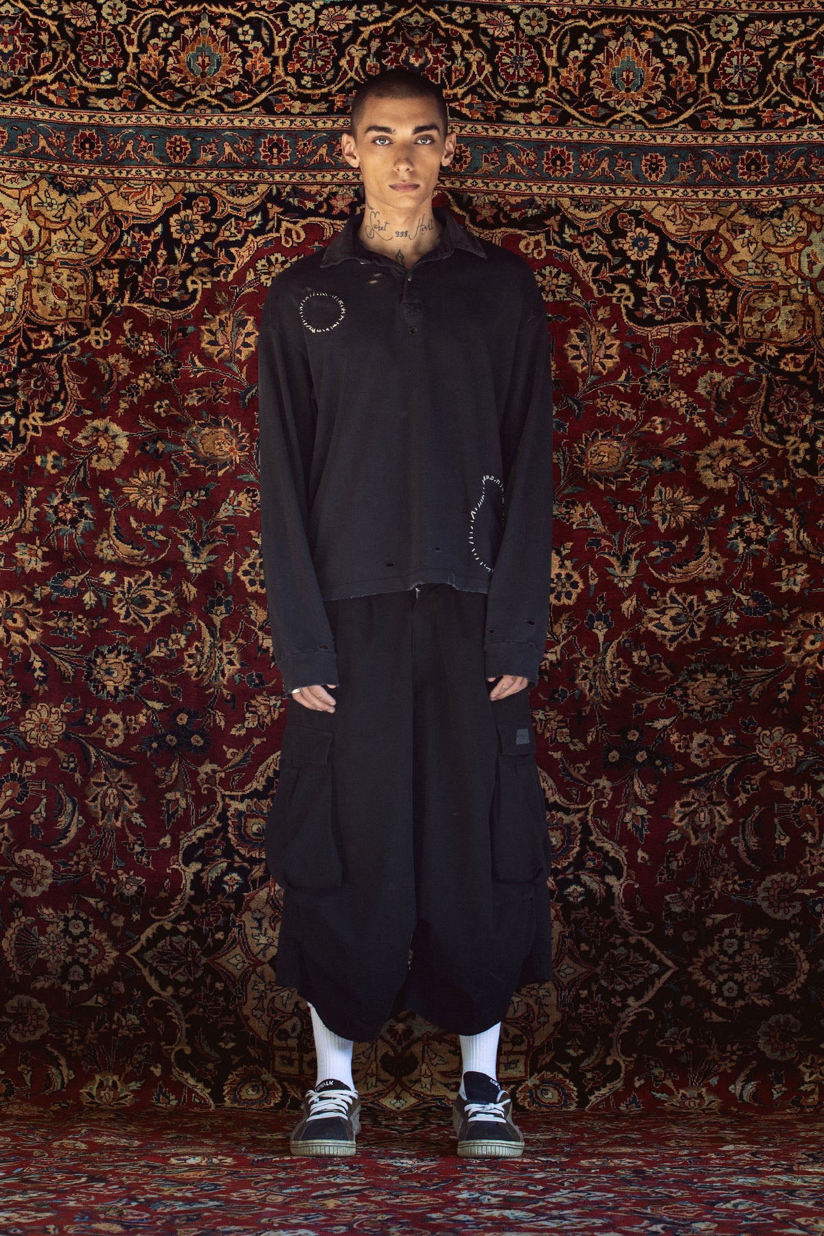 A model stands in front of a large, ornate red and gold patterned rug wearing a black oversized zip-up jacket with circular stitched details on the chest, matching black baggy pants, white socks, and black sneakers.