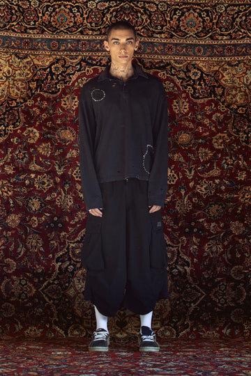 A model stands in front of a large, ornate red and gold patterned rug wearing a black oversized zip-up jacket with circular stitched details on the chest, matching black baggy pants, white socks, and black sneakers.