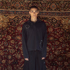 A model stands in front of a large, ornate red and gold patterned rug wearing a black oversized zip-up jacket with circular stitched details on the chest, matching black baggy pants, white socks, and black sneakers.
