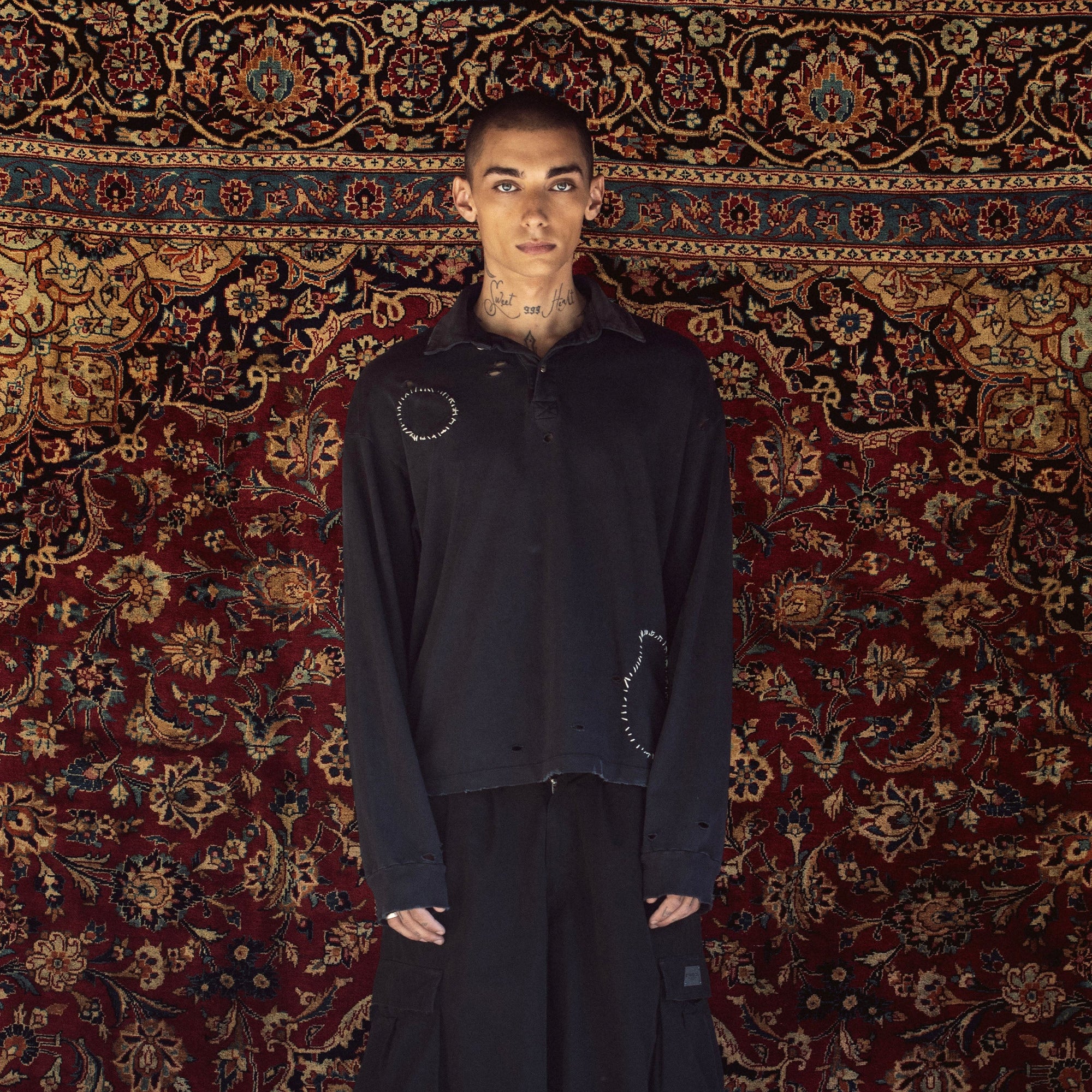 A model stands in front of a large, ornate red and gold patterned rug wearing a black oversized zip-up jacket with circular stitched details on the chest, matching black baggy pants, white socks, and black sneakers.
