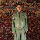 A model stands facing forward wearing a matching light green denim work jacket and wide-leg work pants set, posed against a large, ornate red and gold patterned oriental rug.