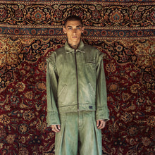 A model stands facing forward wearing a matching light green denim work jacket and wide-leg work pants set, posed against a large, ornate red and gold patterned oriental rug.