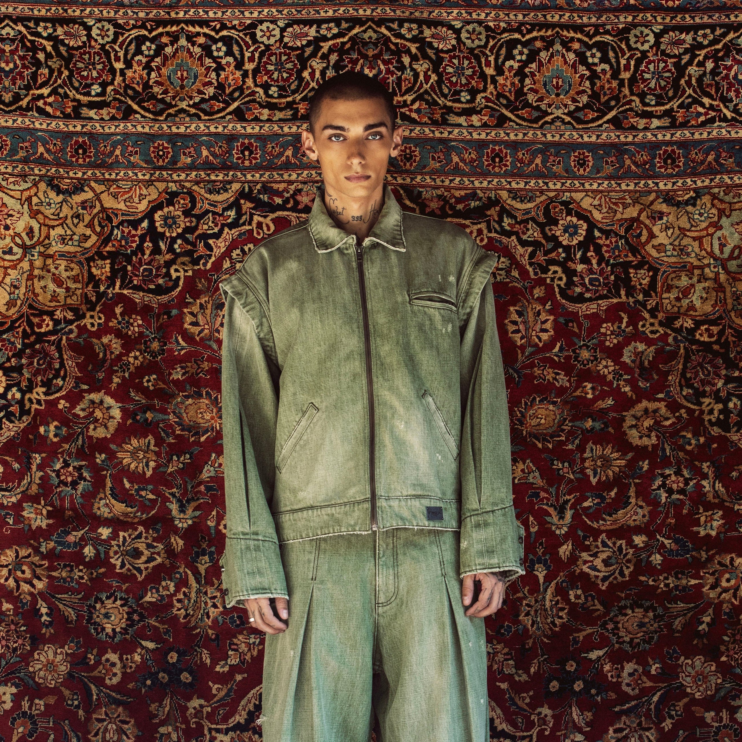 A model stands facing forward wearing a matching light green denim work jacket and wide-leg work pants set, posed against a large, ornate red and gold patterned oriental rug.