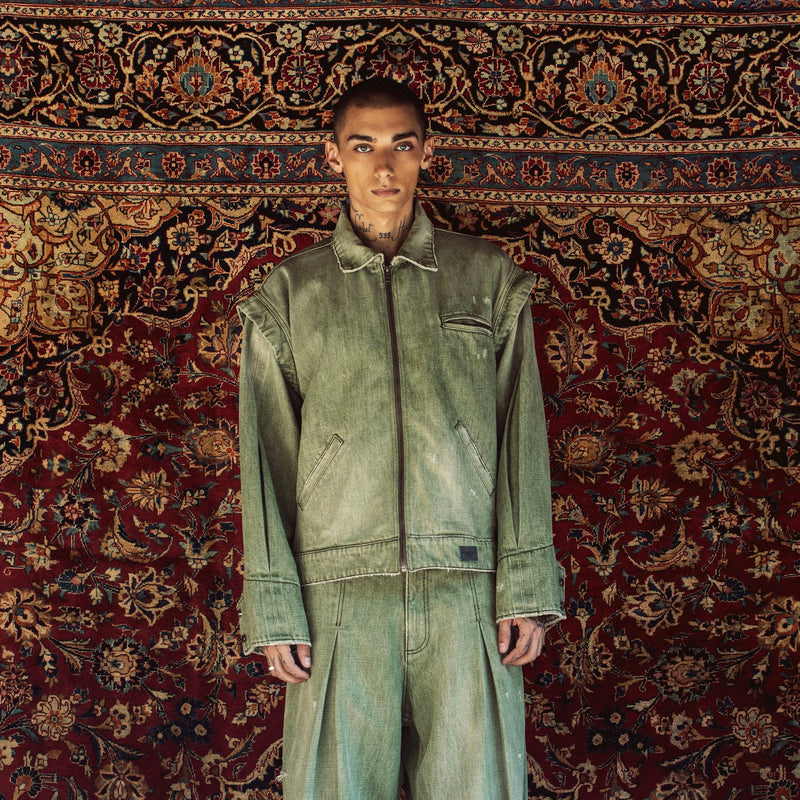 A model stands facing forward wearing a matching light green denim work jacket and wide-leg work pants set, posed against a large, ornate red and gold patterned oriental rug.