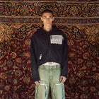 A model stands in front of a large, ornate red and gold patterned rug, wearing a black cropped zip-up hoodie with a white patch on the chest, light-wash distressed green jeans with ripped details, and brown shoes.