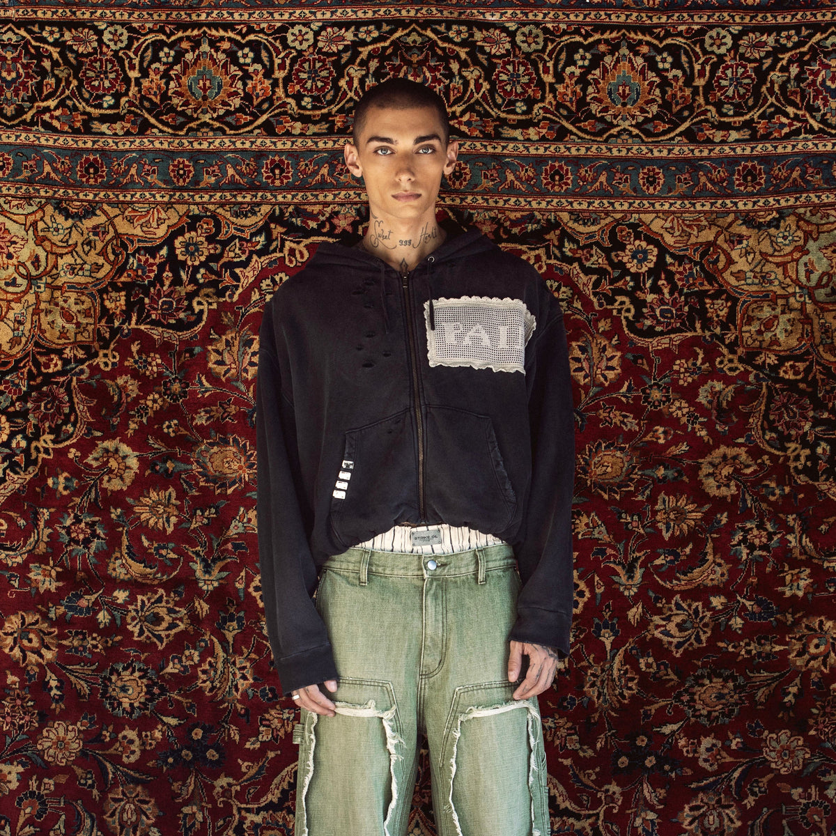 A model stands in front of a large, ornate red and gold patterned rug, wearing a black cropped zip-up hoodie with a white patch on the chest, light-wash distressed green jeans with ripped details, and brown shoes.