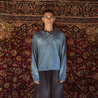 A model stands against a large, ornate red and gold patterned rug, wearing a distressed slate blue long-sleeve polo shirt, dark gray cropped pants, white socks, and black sneakers.