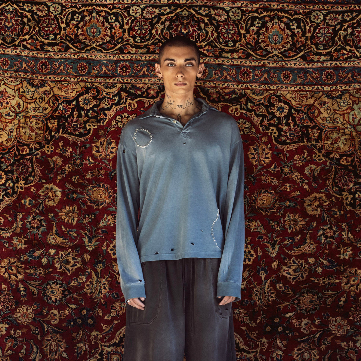 A model stands against a large, ornate red and gold patterned rug, wearing a distressed slate blue long-sleeve polo shirt, dark gray cropped pants, white socks, and black sneakers.