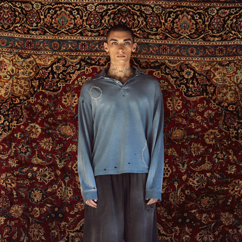 A model stands against a large, ornate red and gold patterned rug, wearing a distressed slate blue long-sleeve polo shirt, dark gray cropped pants, white socks, and black sneakers.