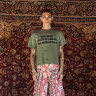 A model stands in front of a large, ornate red and gold patterned rug, wearing a distressed olive green t-shirt with black text and red-and-white patterned wide-leg shorts.