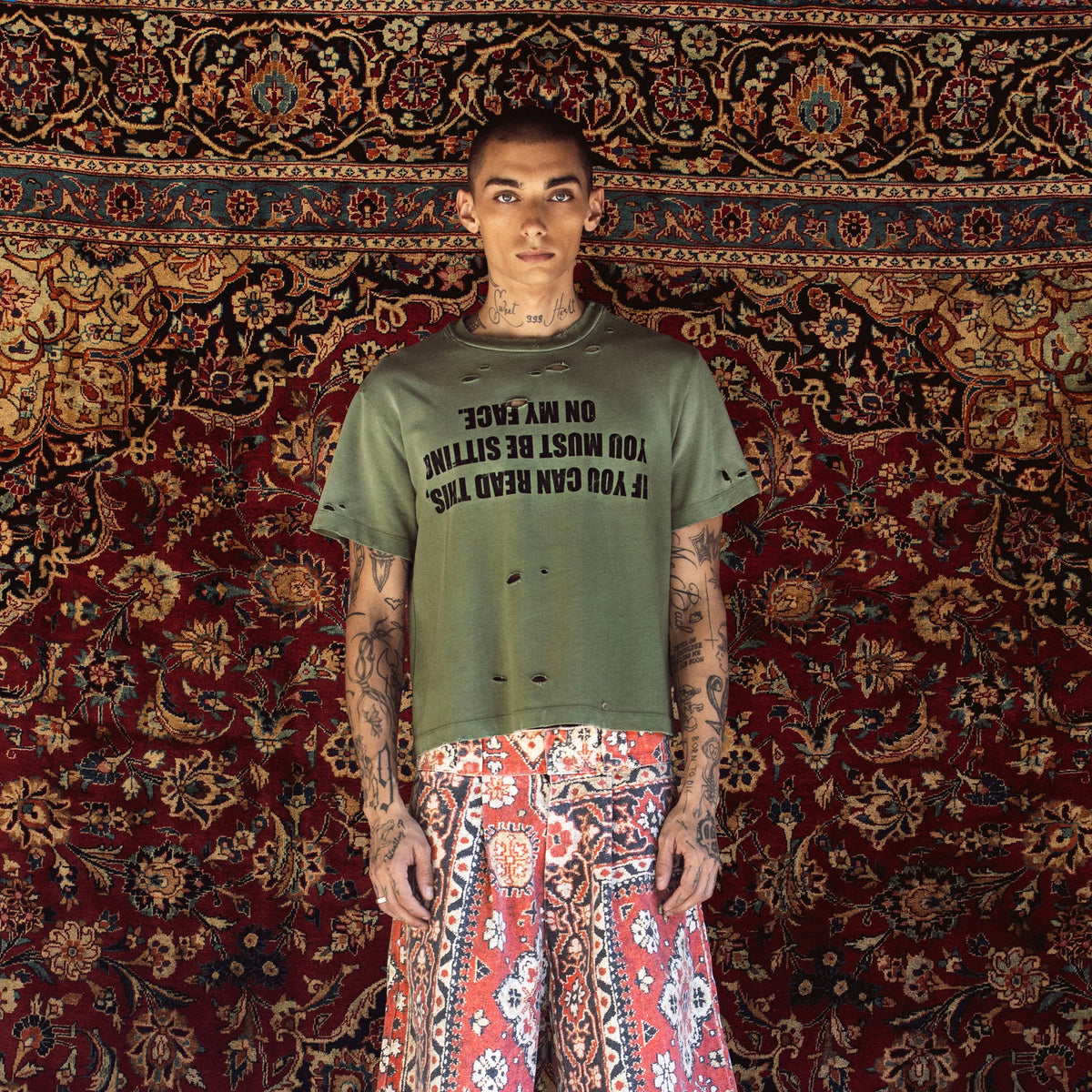 A model stands in front of a large, ornate red and gold patterned rug, wearing a distressed olive green t-shirt with black text and red-and-white patterned wide-leg shorts.