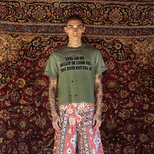 A model stands in front of a large, ornate red and gold patterned rug, wearing a distressed olive green t-shirt with black text and red-and-white patterned wide-leg shorts.