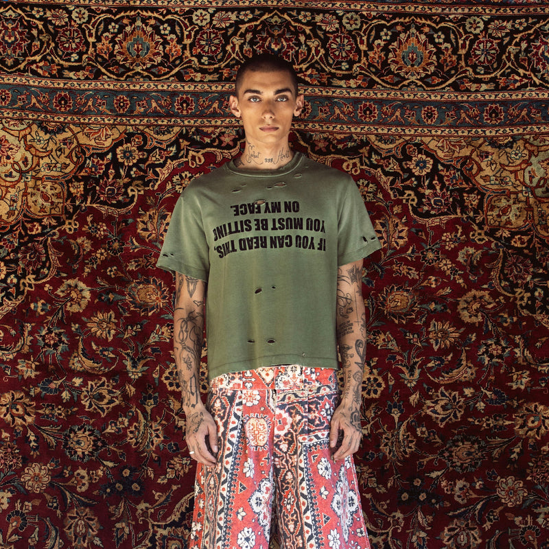 A model stands in front of a large, ornate red and gold patterned rug, wearing a distressed olive green t-shirt with black text and red-and-white patterned wide-leg shorts.